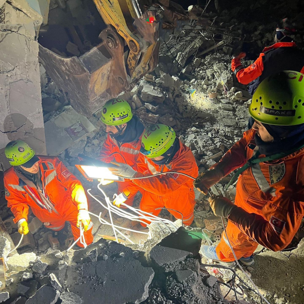 Gruppe von Menschen in orangefarbenen Uniformen und grünen Helmen bei der Arbeit an Trümmern eines eingestürzten Gebäudes mit einem Kran im Hintergrund.