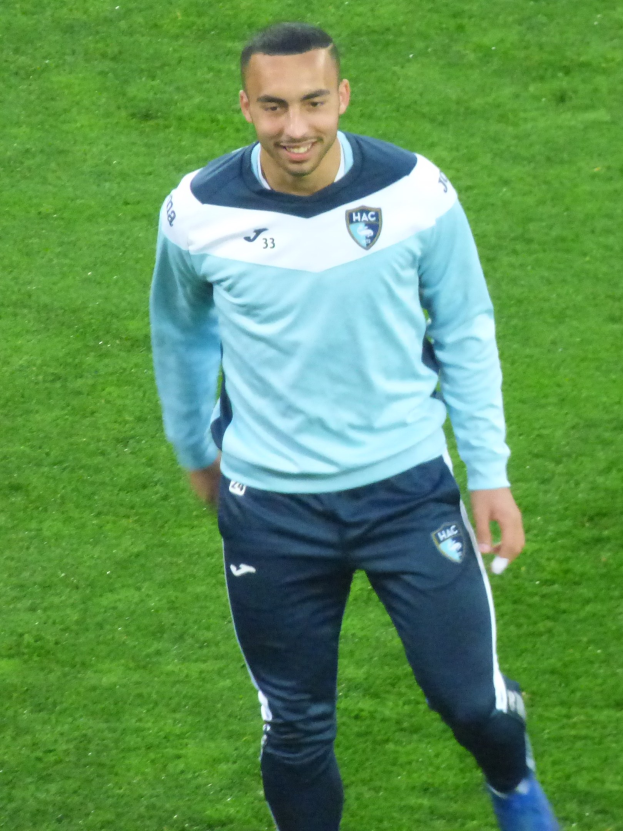 Ein Mann in einem blauen Hemd und schwarzen Hosen läuft auf einem Fußballfeld, lächelt und trägt Sportschuhe.