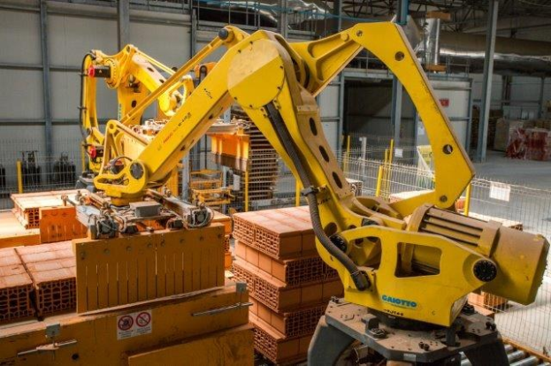 Large industrial warehouse with stacked boxes, overhead cranes, fencing, poles, and a distant wall in an automated manufacturing setting.