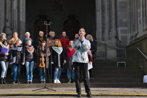 Eine Frau in der Mitte hält ein Mikrofon und singt, umgeben von einer Gruppe von Menschen mit Musikinstrumenten, vor einem Gebäude mit Treppen, Geländern und einer Wand mit Säulen und einer Tür.