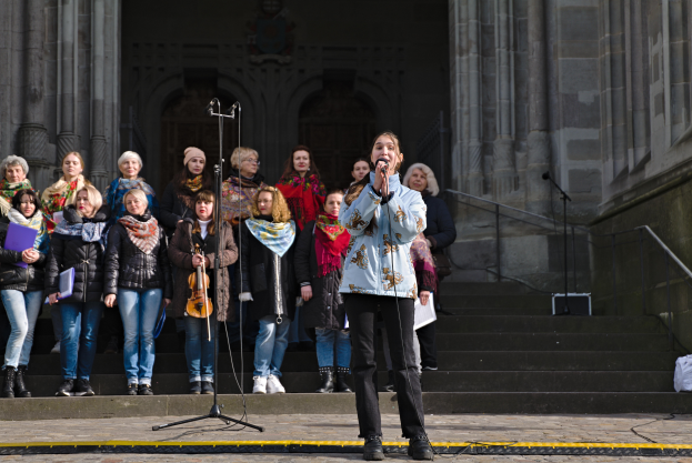 Eine Frau in der Mitte hält ein Mikrofon und singt, umgeben von einer Gruppe von Menschen mit Musikinstrumenten, vor einem Gebäude mit Treppen, Geländern und einer Wand mit Säulen und einer Tür.