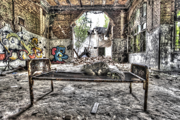 Ein altes Krankenhausbett in der Mitte eines verlassenen Raums mit Graffiti an den Wänden, flankiert von Fenstern auf beiden Seiten und verstreuter Trümmer und Bäumen im Hintergrund.