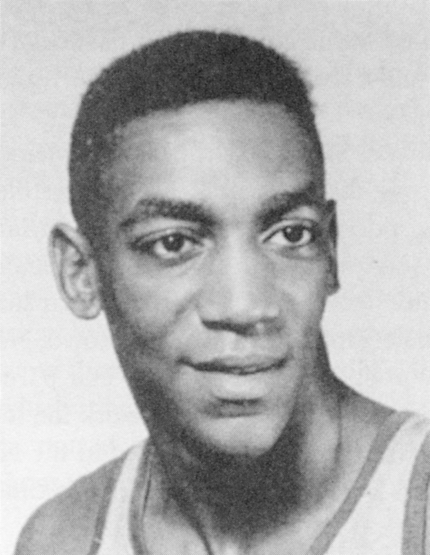 Schwarzes und weißes Foto eines grinsenden Mannes in einem weißen Basketballtrikot mit "Basketball Hall of Fame" darauf, mit zurückgekämmtem Haar und einem Ausdruck von Selbstvertrauen.