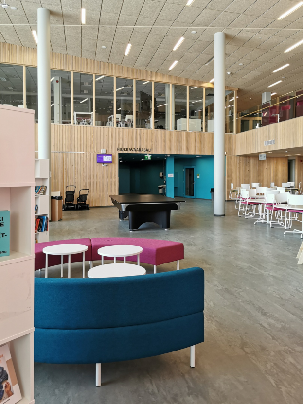 Large room with multiple tables, chairs, sofas, stools, bookshelves, pillars, ceiling lights, and a wall-mounted television, resembling a school library interior.