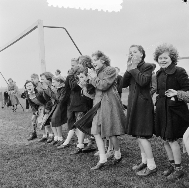 Schwarzes und weißes Bild einer Gruppe von Kindern, die auf einem Feld mit einem Torpfosten und einem klaren Himmel im Hintergrund klatschen.