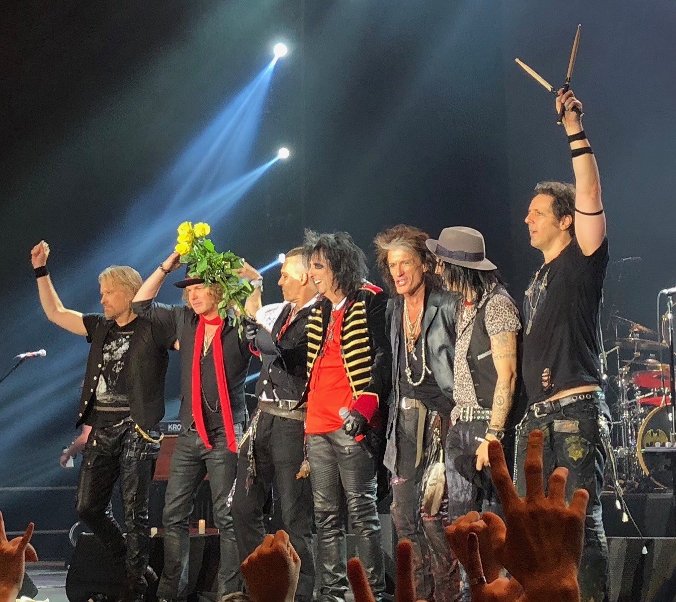 Group of people on a brightly lit stage holding musical instruments, microphones, and a bouquet, with audience hands visible at the bottom.