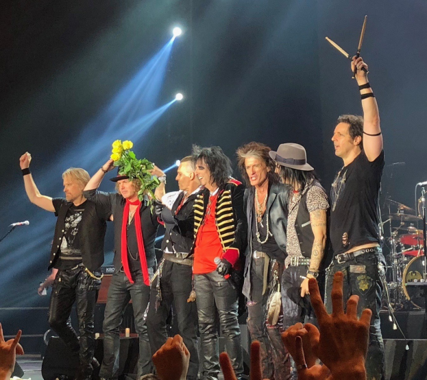Group of people on a brightly lit stage holding musical instruments, microphones, and a bouquet, with audience hands visible at the bottom.