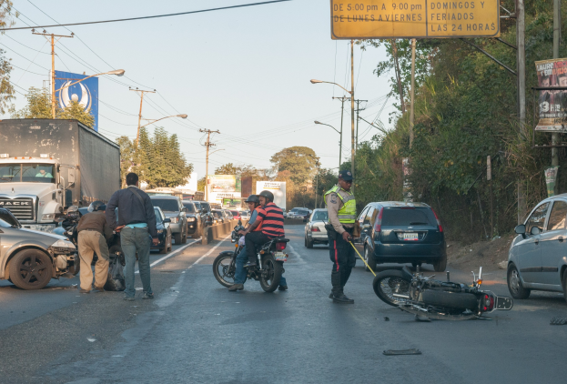 Gruppe von Menschen um ein verunglücktes Motorrad auf der Straßenseite mit mehreren Fahrzeugen, Bäumen, Strommästen und einem klaren Himmel im Hintergrund.