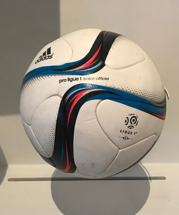 Ein weißer Fußball mit schwarzer 'Ligue 1'-Text und Adidas-Logo liegt auf einem weißen Tisch gegen einen hellen weißen Hintergrund.