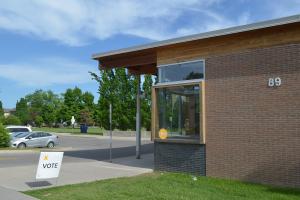 Ein Ziegelgebäude mit einem "Wahl"-Schild davor, umgeben von Gras, Fahrzeugen, Pfählen, Brettern, Bäumen, anderen Gebäuden und einem bewölkten Himmel.