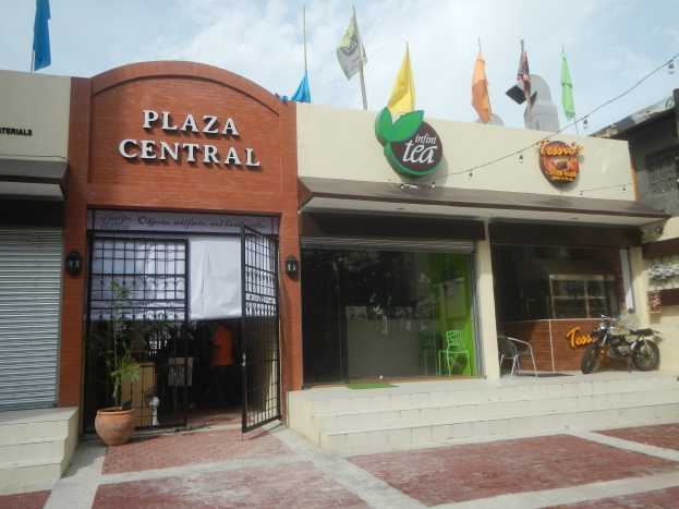 Ein zentraler Platz in Cebu City, Philippinen, mit umliegenden Gebäuden, Motorrädern, Outdoor-Sitzgelegenheiten, Hauspflanzen, Flaggen, Beschilderung, elektrischen Oberlichtern und Kabeln sowie einem bewölkten Himmel.