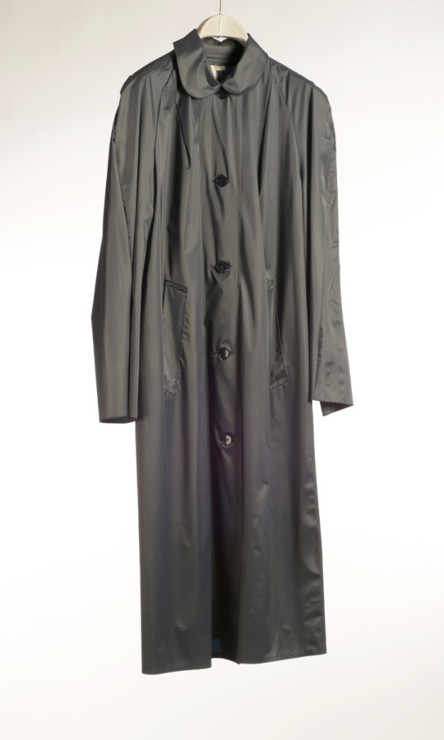 Schwarzer glänzender Trenchcoat mit doppelreihiger Knopfleiste, Kragen, zwei großen Taschen und Reißverschluss, der an einer weißen Wand hängt.