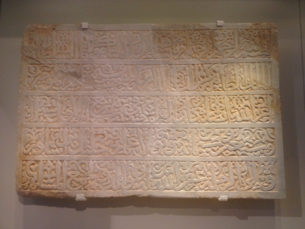 Steintafel mit arabischer Kalligrafie in leuchtenden Farben, die eine detaillierte Schrift und einen dekorativen Rahmen aufweist, auf einer neutralen Hintergrundwand montiert.