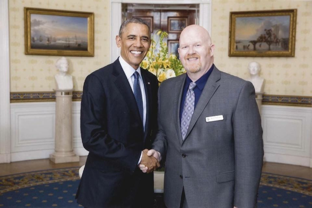 Präsident Obama gibt einem Mann im Anzug die Hand im Oval Office des Weißen Hauses, umgeben von dekorativen Elementen wie einem Teppich, Statuen, einer Blumenvase und gerahmten Fotos an der Wand.