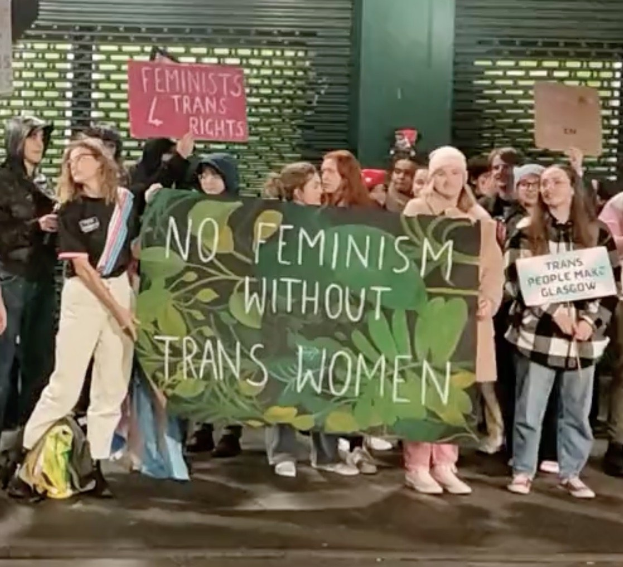 Eine Gruppe von Menschen hält ein Banner mit der Aufschrift 'Kein Feminismus ohne trans Frauen' und Schilder in einem öffentlichen Raum mit einer Wand und einer Rolllade im Hintergrund.