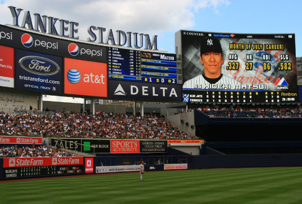 A view of Yankee Stadium with a grass field, a person standing, advertisement boards, seated spectators, and a screen above.