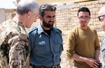 Gruppe von Männern, die vor einer Ziegelwand stehen und sich unterhalten, wobei einer ein Objekt hält und der US-Verteidigungsminister von der irakischen Armee interviewt wird.