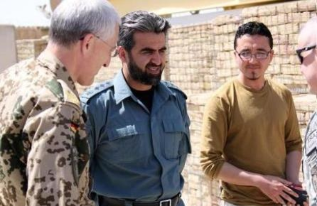 Gruppe von Männern, die vor einer Ziegelwand stehen und sich unterhalten, wobei einer ein Objekt hält und der US-Verteidigungsminister von der irakischen Armee interviewt wird.