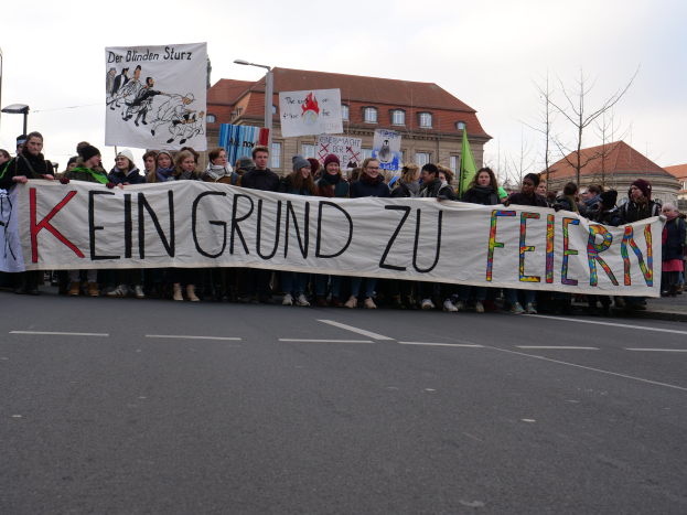 Demonstranten mit einem Transparent "Kein Grund zu Feiern" gegen deutsche Sparmaßnahmen, mit Straßeninfrastruktur und Gebäuden im Hintergrund.