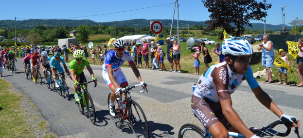 Gruppe von Radfahrern fährt auf einer Straße mit Zuschauern an den Seiten, einige tragen Helme und Schutzbrillen, andere halten Fahnen, vor einer Kulisse aus Bäumen, Schildern, Fahrzeugen, Zelten, Gebäuden, Hügeln und einem klaren blauen Himmel.