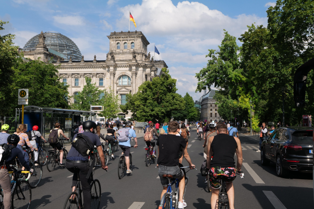 Group of people riding bicycles down a tree-lined street in Berlin, Germany, with buildings and a bus stop visible, under a cloudy sky and a flag flying on a building.
