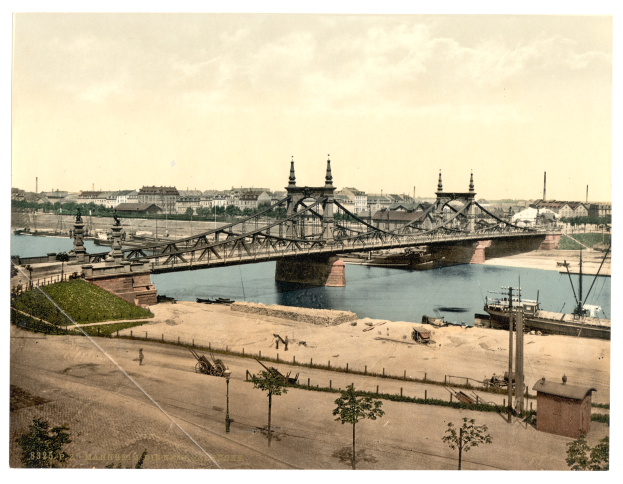 Altes Schwarz-Weiß-Foto einer Brücke über einen Fluss mit Booten darunter, flankiert von grünem Land mit Bäumen und Pfählen auf der linken Seite und einer Skyline von Gebäuden unter einem klaren Himmel im Hintergrund.