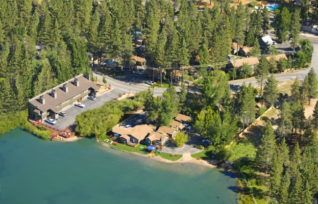 Luftaufnahme eines Resorts am See, umgeben von B├Ąumen, H├Ąusern, Fahrzeugen, Stra├čen, Strommasten und Gras, mit dem See unten sichtbar.