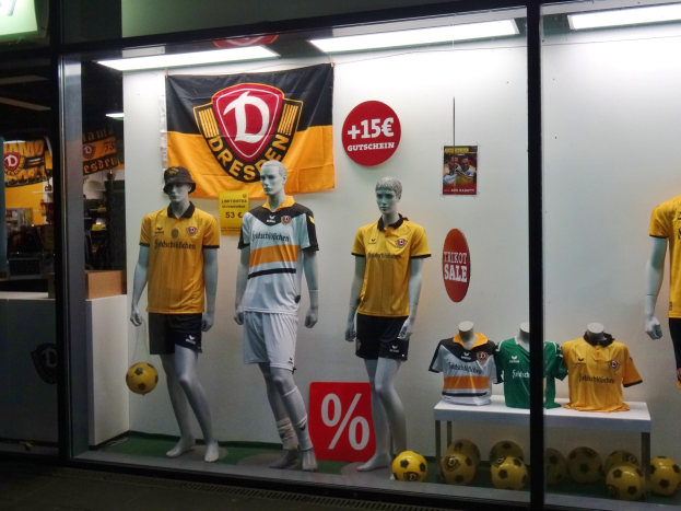 Ladenfenster-Dekoration mit Puppen in gelben und schwarzen Fußballtrikots, einem Tisch mit T-Shirts und Fußballs, einer Banner mit Text und Logo, Deckenbeleuchtung und einer Schiefertafel mit Schrift links.