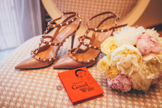 Ein Tisch zeigt ein Buch mit dem Titel 'Wie man eine gute Ehefrau ist', einen Blumenstrauß und ein Paar Valentino-Schuhe, vor einem Vorhang.