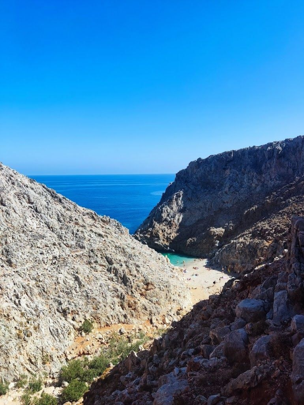 Panoramablick auf einen Mittelmeerstrand von einem Berg aus, mit klarem blauem Wasser, grüner Vegetation, steinigen Hügeln und einem blau-weißen Himmel.