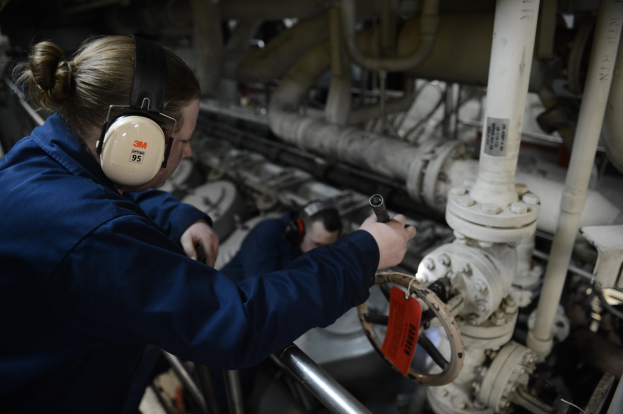 Zwei Frauen in blauen Uniformen arbeiten an einer industriellen Maschine, eine hält ein Objekt und trägt Kopfhörer, mit Rohren und Equipment im Hintergrund.