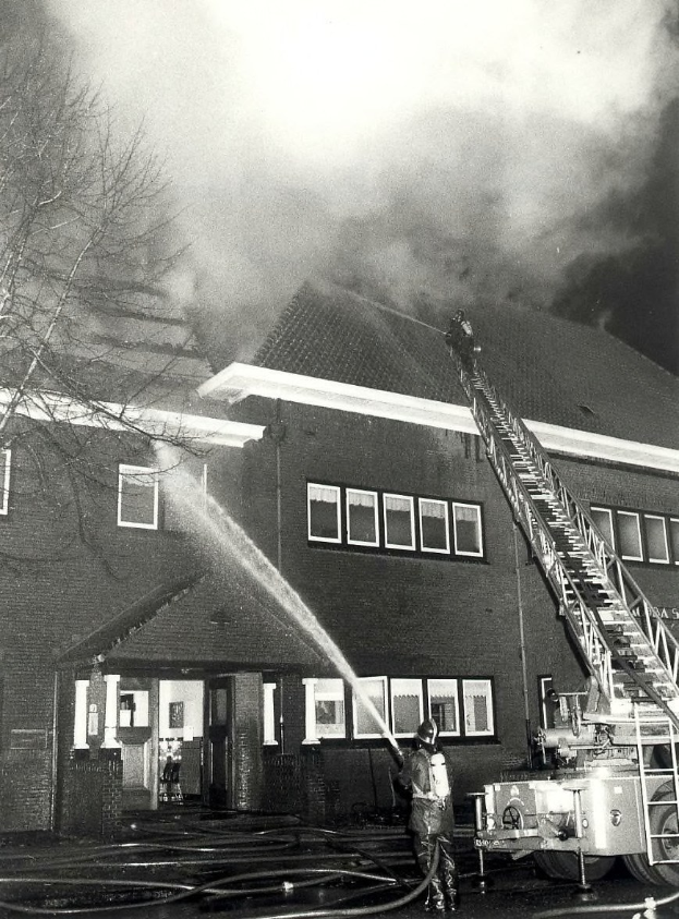 Schwarzes und weißes Foto eines Feuerwehrautos, das Wasser auf ein Gebäude mit Fenstern und Türen sprüht, einen Baum auf der linken Seite und einen bewölkten Himmel im Hintergrund.