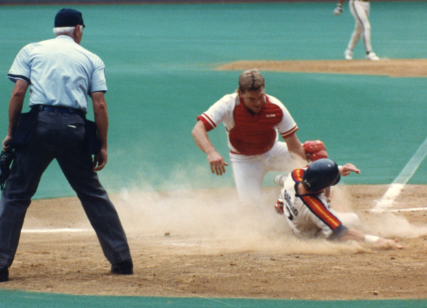 Baseballspieler rutscht auf Home Plate, Umpire steht in der Nähe, Zuschauer und Wand im Hintergrund sichtbar.