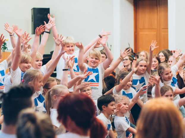 Gruppe von Kindern, die vor einer Menge die Hände heben, mit einem Tisch, einer Vase, einem Lautsprecher, einer Wand und einer Tür im Hintergrund.