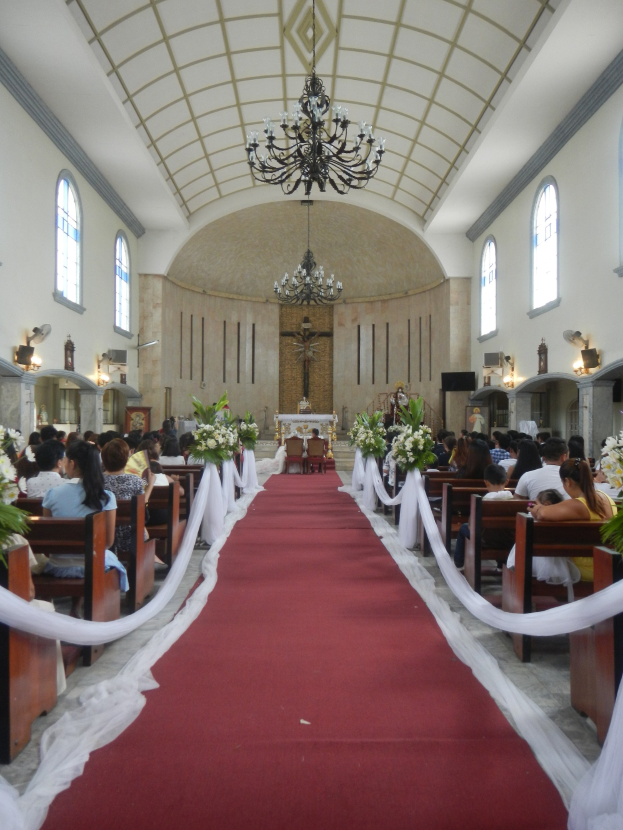Hochzeitszeremonie in einer Kirche mit einem roten Teppichgang, der mit Blumenbouquets und weißen Tüchern gesäumt ist, flankiert von Bänken und einem Kreuzsymbol an der Wand im Hintergrund, mit zwei Kronleuchtern, die von der Decke hängen.