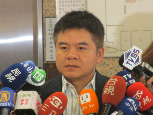 Ein Mann im Anzug spricht vor einer wandmontierten Tafel mit Reportern, waehrend eine Frau rechts eine Kamera halt.