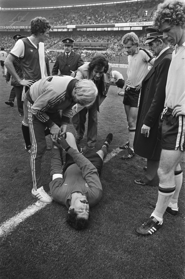 Schwarzes und weißes Bild eines Fußballspielers, der auf dem Boden liegt, während eine Gruppe von Männern und eine Stadionmenge um ihn herumsteht, was darauf hindeutet, dass er möglicherweise verletzt ist.