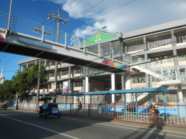 Eine belebte Stadtstraße mit Fahrzeugen, eine Brücke mit Geländern und Treppen, Strommasten mit Drähten, Laternen, Bäume, Gebäude mit Fenstern, Banner mit Text und ein bewölkter Himmel.
