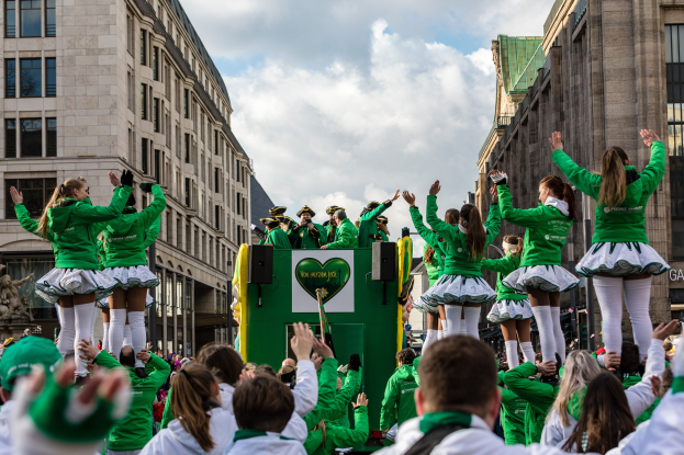 Gruppe von Menschen in grünen Hemden und weißen Röcken auf einem geschmückten Festwagen bei einem St. Patrick's Day Umzug, mit Gebäuden, einer Statue und einem klaren blauen Himmel im Hintergrund.