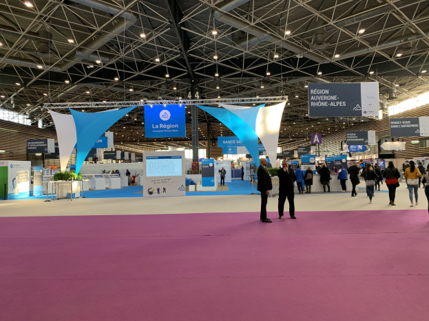 A large trade show booth with numerous attendees walking around, some carrying bags, under illuminated ceiling banners and tables displaying various items.