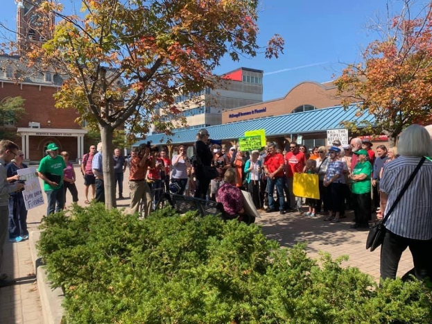 Eine Gruppe von Menschen demonstriert vor einem Gebäude auf dem Campus der University of Michigan, mit Schildern und einigen mit Kameras, Taschen und Kappen, mit Bäumen und Himmel im Hintergrund.