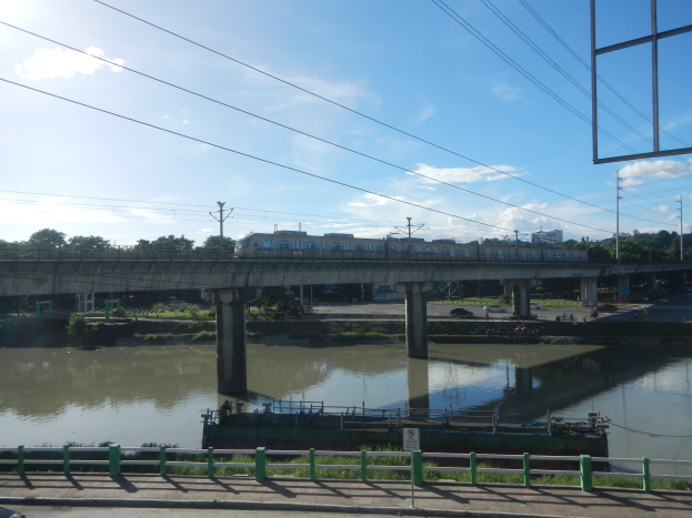 Ein Zug überquert eine Brücke über einen Fluss, mit Fahrzeugen auf der Straße darunter, einem Flussufergeländer, Bäumen im Hintergrund, Strommasten mit Drähten und einem bewölkten Himmel.