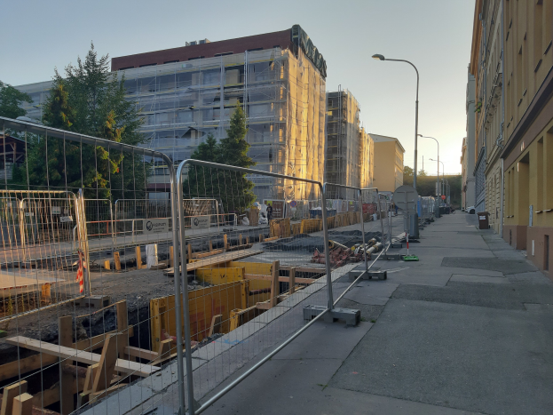 Stadtstraße mit einer Baustelle in der Mitte, umgeben von Gebäuden, Straßeninfrastruktur, Fahrzeugen, Bäumen und einem klaren blauen Himmel.