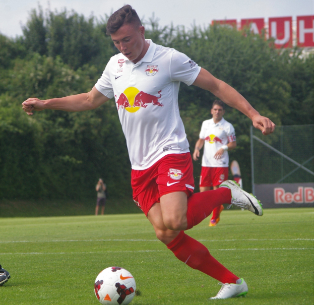 Ein professioneller Fußballspieler, der einen Ball auf einem Rasenfeld kickt, mit "RB Leipzig" im Hintergrund und einem klaren blauen Himmel.