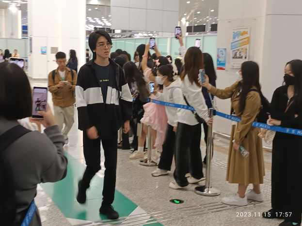 People standing in line at an airport with barrier poles, ribbons, posters on walls, and ceiling lights, with a watermark indicating the Seoul International Airport.
