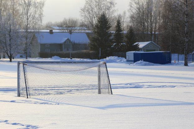 Fussballtor auf einem verschneiten Feld mit Netz, Pfosten, einem Zaun, einem Container, Bäumen, Häusern und einem bewölkten Himmel.