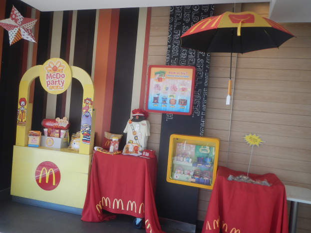 Ein McDonald's thematisierter Geburtstagstisch mit einer roten Decke und einem Schirm, der verschiedene Gegenstände zeigt, vor einer Wand mit einer Tafel, unter einer sichtbaren Decke.