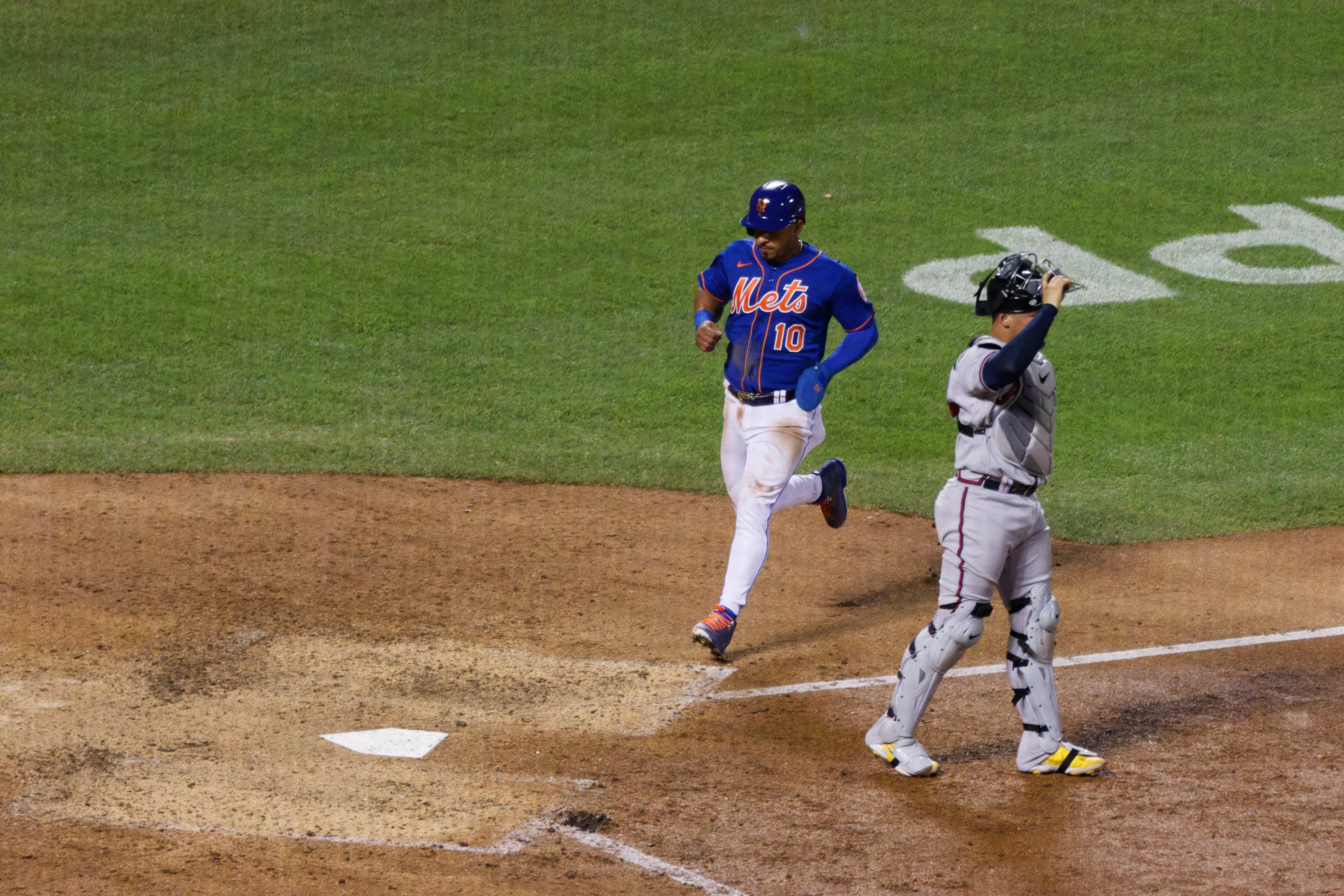 Baseballspieler in Uniform und Helm rennt Richtung Home Plate und hält einen Schläger in der Hand auf einem grasbewachsenen Feld mit Text am Boden.