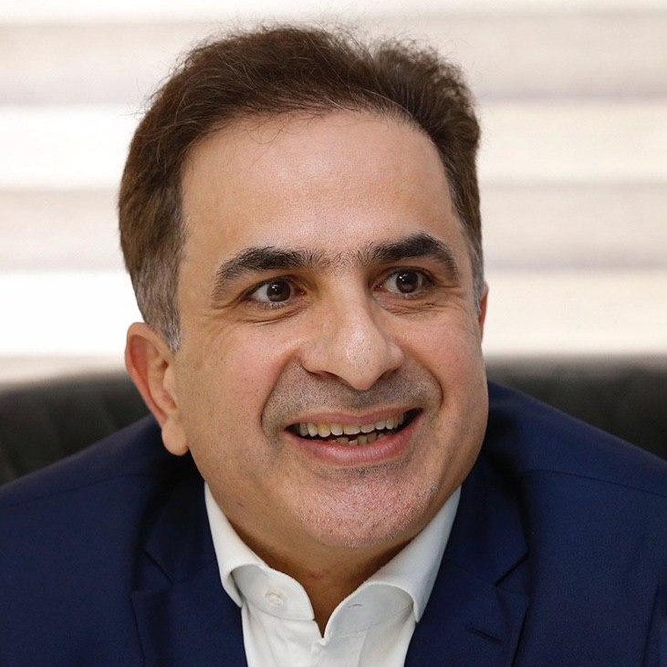 Ein Mann in einem blauen Anzug, der strahlend in die Kamera lächelt, vor einem unscharfen Hintergrund.