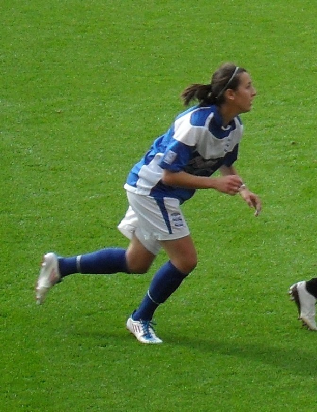 Eine Fußballspielerin in einem blauen und weißen Trikot, die auf einem Feld läuft, wobei ein Teil des Beins eines anderen Spielers rechts zu sehen ist.